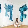 Kayta is sitting on the floor in a corner of a room. Behind her are three of her figurative paintings hung from ceiling to floor on the wall.
