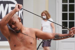 Luis is wrapping a braided cord loop around his head and neck while his arms stretch the cord loop to the right. His eyes are closed as he caresses his head into the crook of his left elbow.