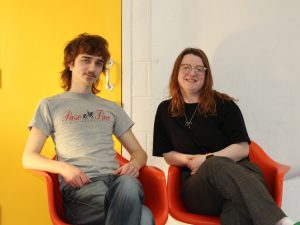 two people sitting in red chairs beside each other in front of a white wall
