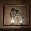 Two gas lamps are stretched out and leaning on one another. They are within a metal box with the sides taken out. They are glowing in a dimly lit room.
