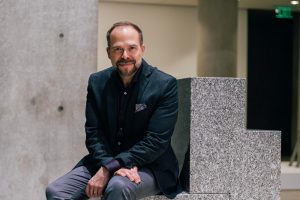 David is still on the left side of a geometric stone sculpture. He is wearing a suit and looking directly at the camera with a slight smile. His hands are in his lap, and his ankles are crossed.