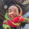 Hannah is sitting outside by the exterior of a house. The camera is close to her face, and Hannah is looking down at it, smiling. Only her head and shoulders are visible. Her head is wrapped in a bright green and pink scarf.