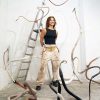 Sara is standing in a corner of a room and in front of a step ladder with her hands behind her back and smiling at the camera. Around her on the floor and the walls are her sculptures: long tubes that resemble large earthworms. The sculptures are painted white, brown, and black.