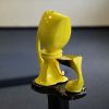 A small, yellow ceramic structure is on a small, black pedestal. The yellow sculpture is made of two parts. The one on the left is taller with a bulbous tip, a handle, and a spout. The spout had a small flame at the end. The flame is under the right part of the sculpture, which is in a birdbath shape and is holding a liquid.