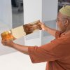 Lorna is holding a strip of golden sheet metal in front of her. The reflection in the sheet metal distorts and stretches Lorna's face.