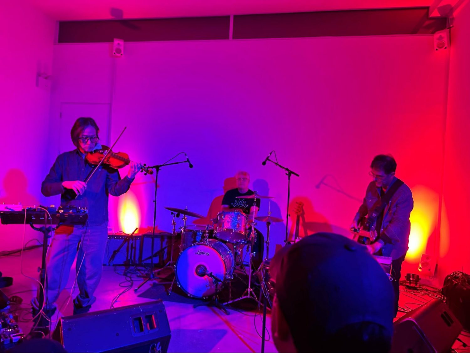 Stephen Vitiello, Brendan Canty, and Hahn Rowe on stage performing. The set is bathed in purple and blue light, with small orange and yellow spotlights on a plain background wall to add harsh visual contrast to the mood of the stage. 