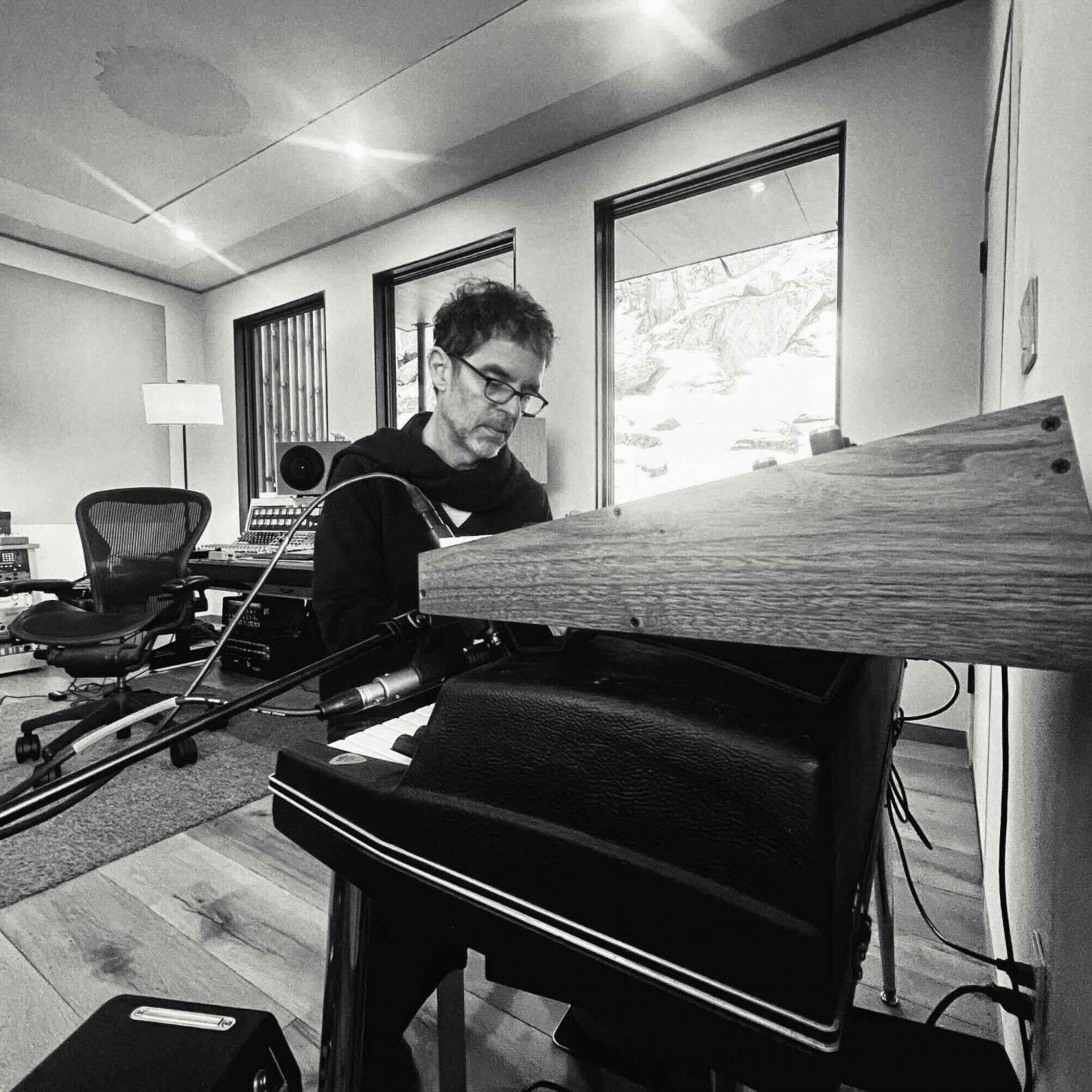 A black and white photograph of Stephen Vitiello working within a studio, sitting at an electric piano and soundboard setup.