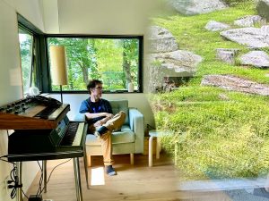 Stephen Vitiello sitting in a spring green living room, with electronic keyboard and soundboard equipment in view. Stephen looks to the viewer's right, where the reflection of a vibrant green hillside dotted with rocks captured in glass is superimposed over the right side of the room.