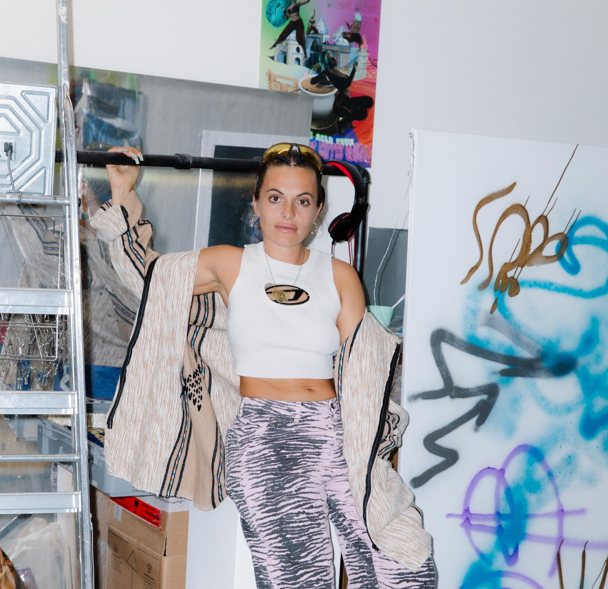 Anna is leaning against a wall, holding onto a pipe that is slightly above her head. Behind her are fragments of her artwork and storage.