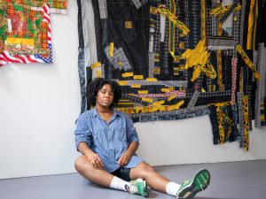 Kat Thompson, wearing a blue shirt, sits on the floor in front of her artwork