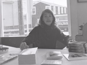 In a black and white photo, a young Mary Oliver sits at a desk surrounded by papers and books
