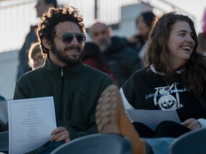 Two students, holding pieces of paper, sit in stadium seats with people around them.