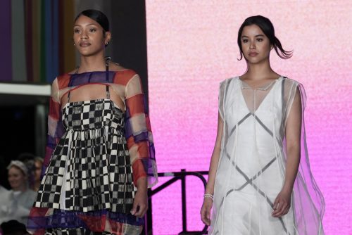 two models walking down the runway. the model on the left is wearing a multicolor patterned dress, while the model on the right is wearing a white and grey dress