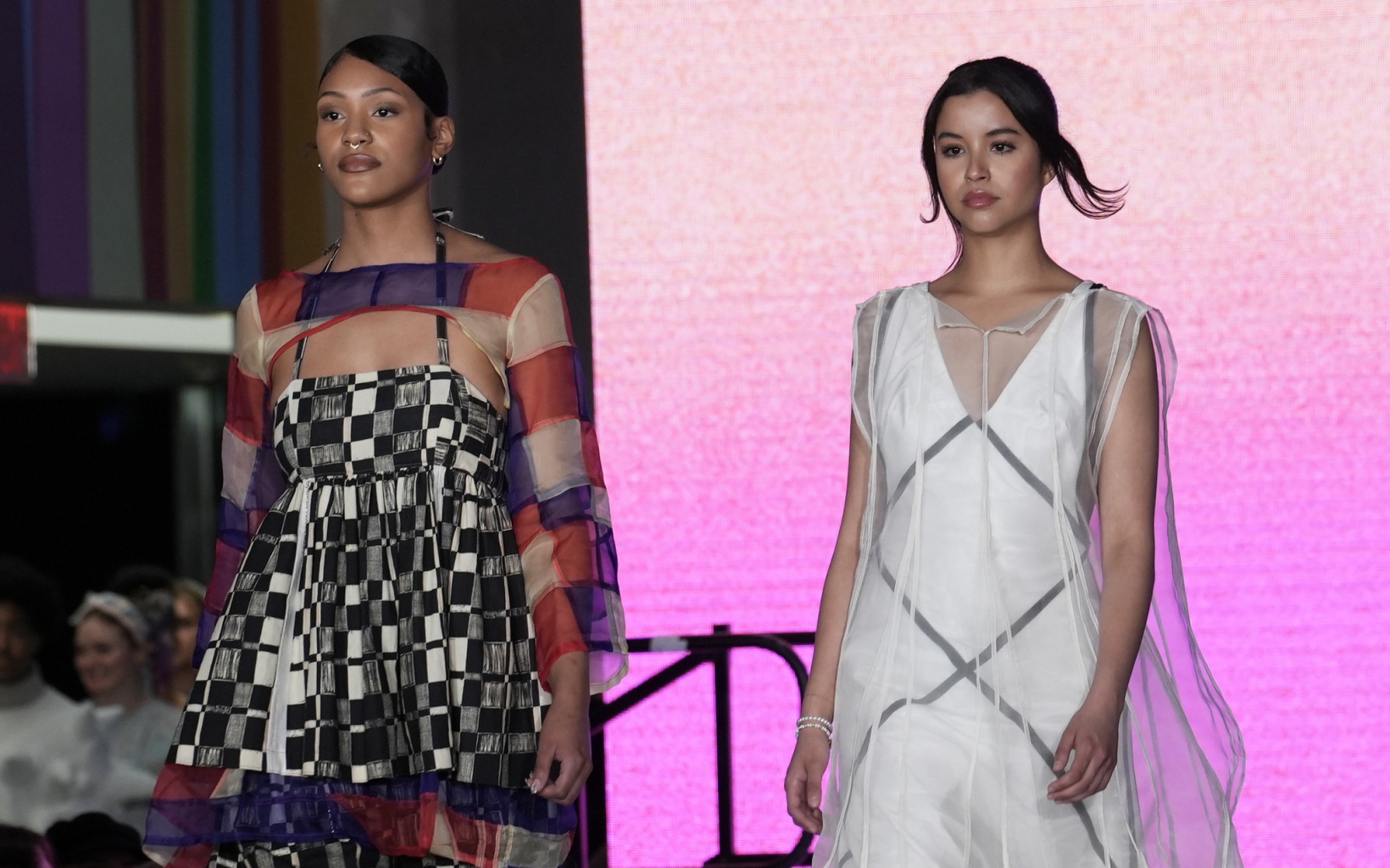 two models walking down the runway. the model on the left is wearing a multicolor patterned dress, while the model on the right is wearing a white and grey dress