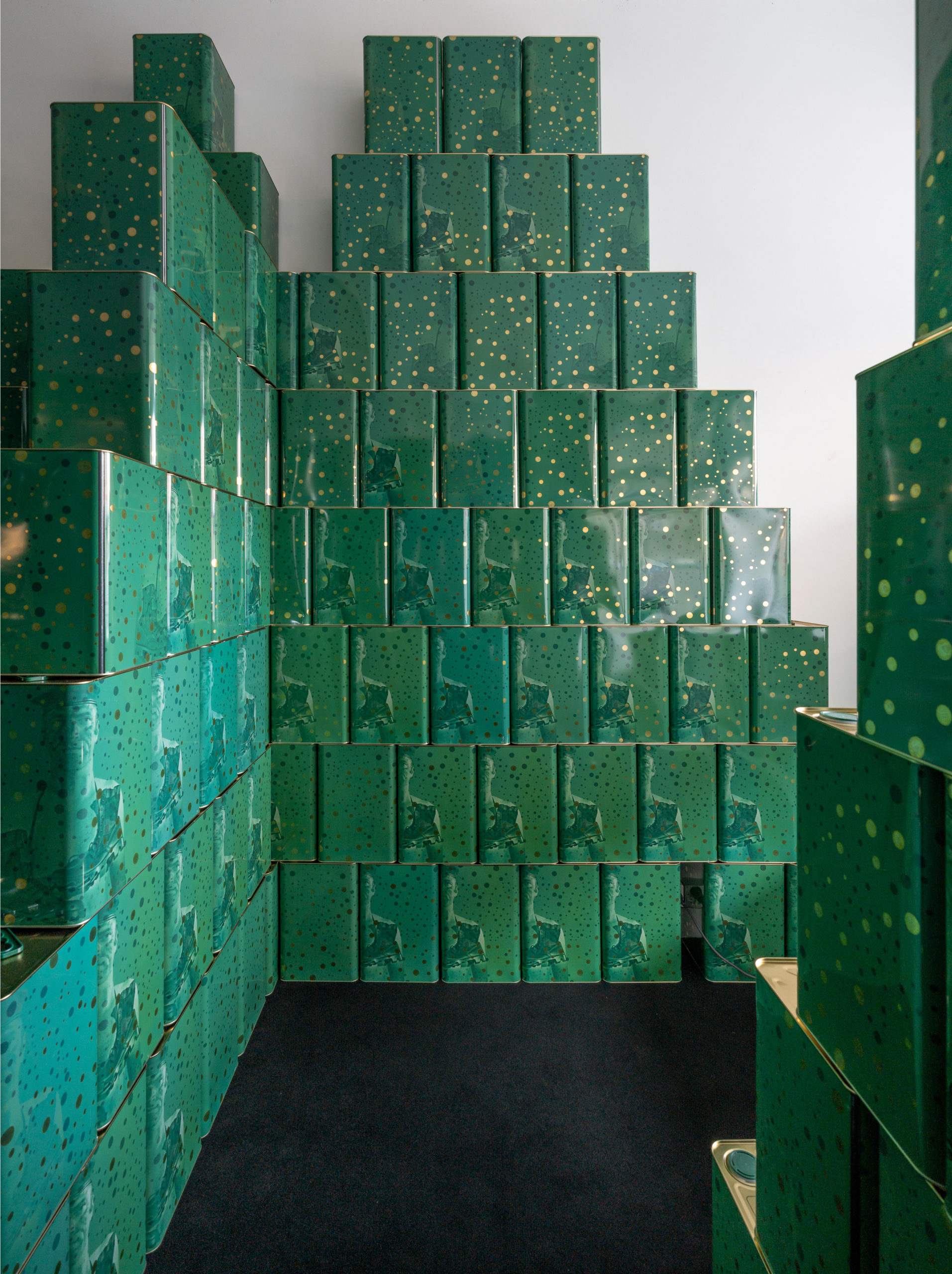 A sculpture made of rectangular green metal boxes which together form tall walls. Each metal box has golden dots. Some metal boxes also have a face of a man with boots tied around his neck.