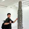 A man wearing a backpack stands and touched part of a tall concrete sculpture with rope tied around it.