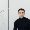 A man stands next to part of a sculpture with has a loop of wire with a star-shapped soft sculpture hanging off of the wire.