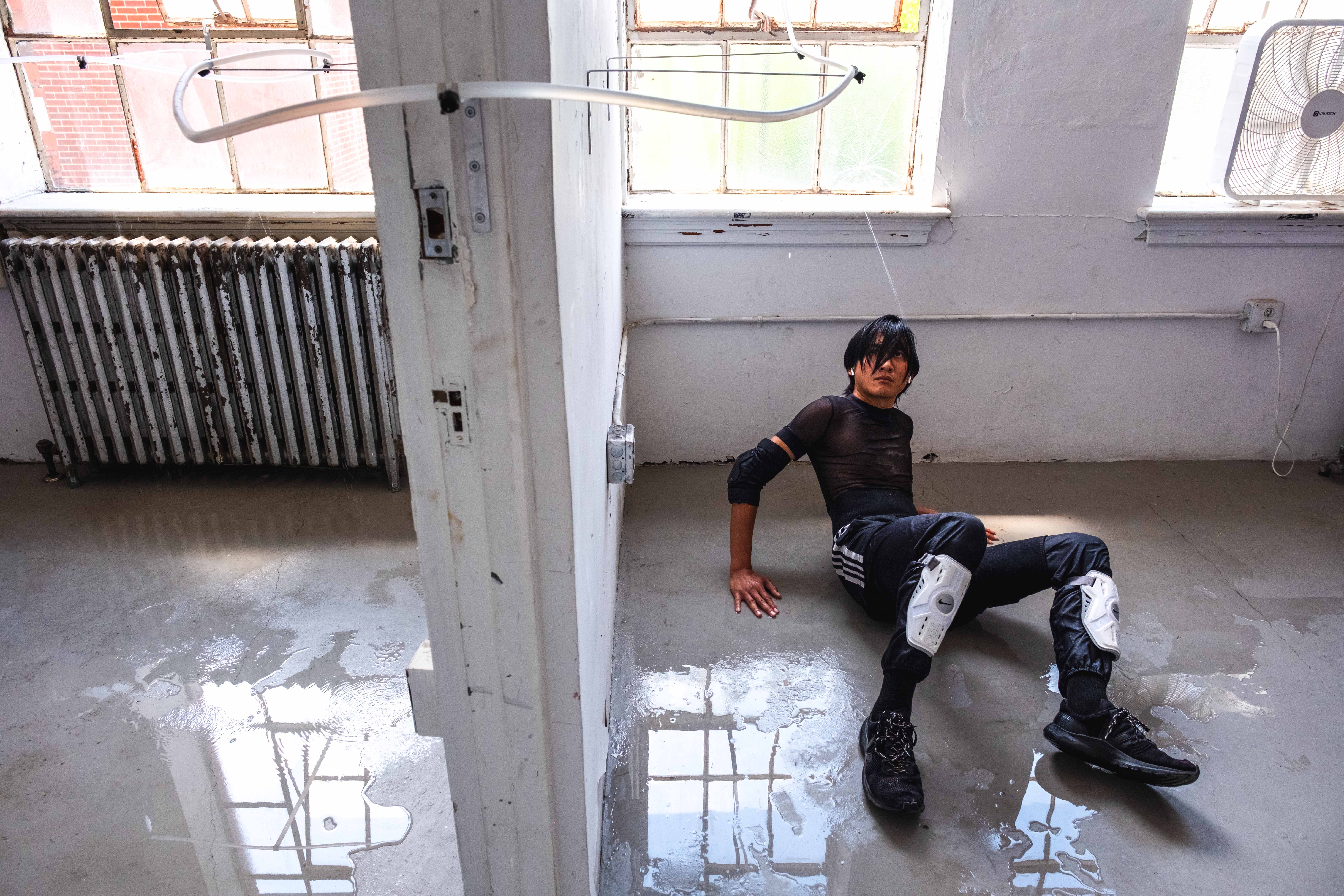 A person wearing elbow pad and shin guards sits on the ground reclining back. They are looking at a leaking pipe that is going around the room.